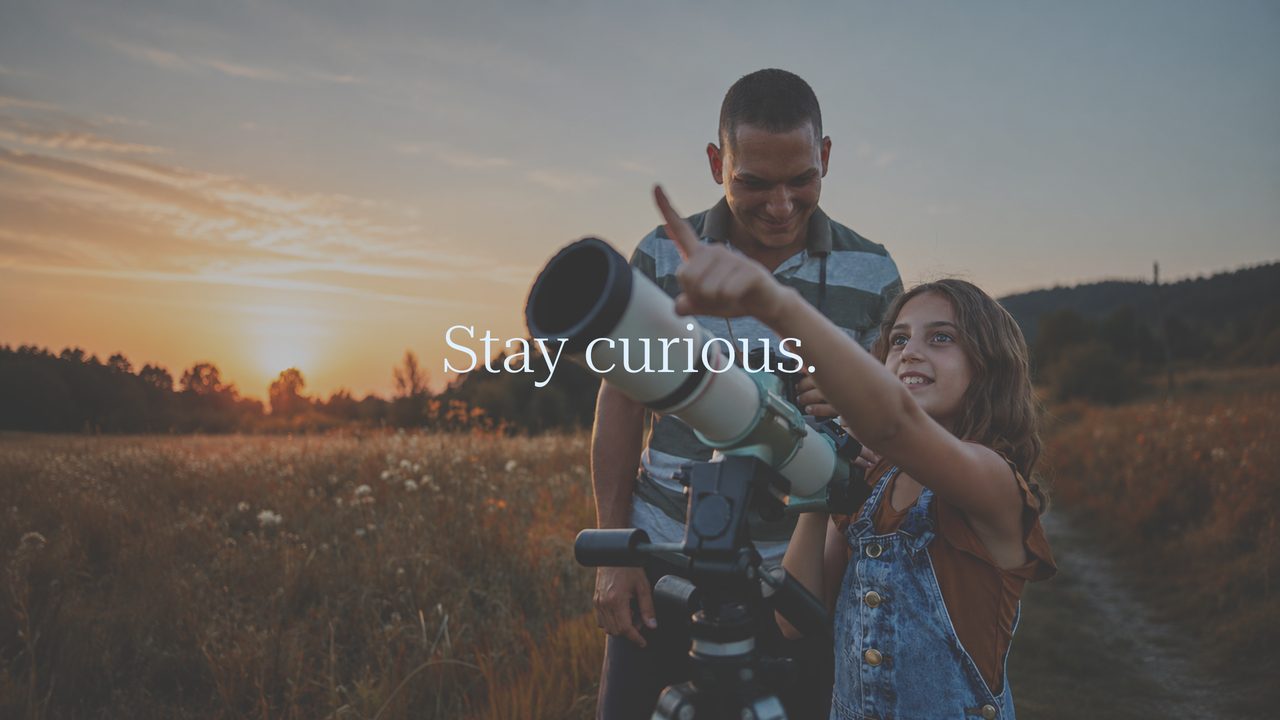 A father and his daughter beside a telescope in a field at sunset, she points at the sky. Across the image: Stay curious.